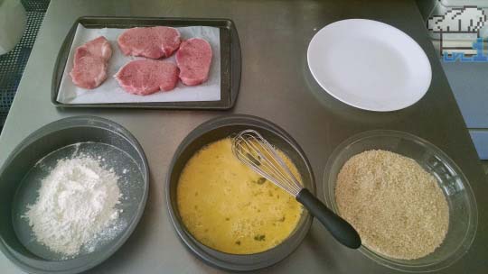 A breading station set up for pork chops, consisting of flour, beaten egg, and breadcrumbs.